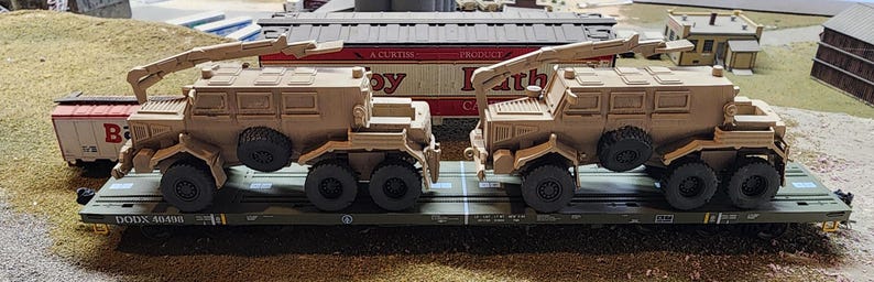 May include: Two tan military vehicles with cranes are being transported on a flatbed train car. The vehicles have a boxy design with a large cab and a flatbed in the back. The train car is labeled "DACOXX 4000".