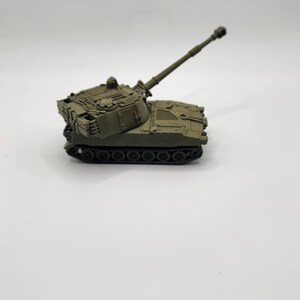 May include: A green toy model of a self-propelled howitzer artillery piece with a long barrel and a rotating turret.