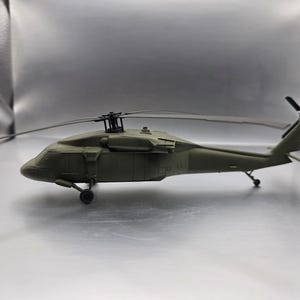 Puede incluir: Un modelo de helicóptero verde oliva con un diseño detallado. El helicóptero de juguete tiene un rotor principal, un rotor de cola y un tren de aterrizaje. El modelo está sobre un fondo neutro.