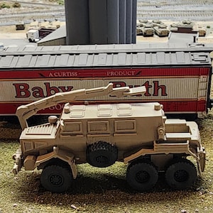 May include: A tan military vehicle with a crane arm, a large box bed, and six wheels. The vehicle is parked next to a red and white boxcar with the words "A Curtis Baby Go-Kart Path" printed on the side.