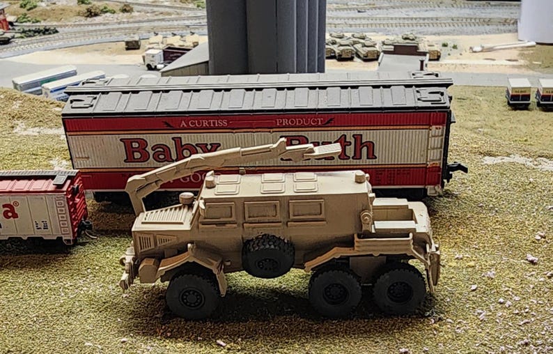 May include: A tan military vehicle with a crane arm, a large box bed, and six wheels. The vehicle is parked next to a red and white boxcar with the words "A Curtis Baby Go-Kart Path" printed on the side.