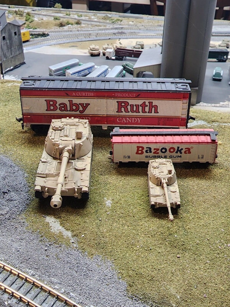 May include: A model train set with a red boxcar with the words "Baby Ruth Candy" on the side, a beige tank, and a smaller beige tank with a red boxcar with the words "Bazooka Bubble Gum" on the side.
