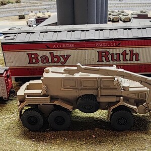 May include: A tan toy military vehicle with a crane arm, a red and white toy boxcar with the text "A Curtiss Product Baby Ruth" on the side, and a red toy boxcar with the text "Booka Bubble Gum" on the side.