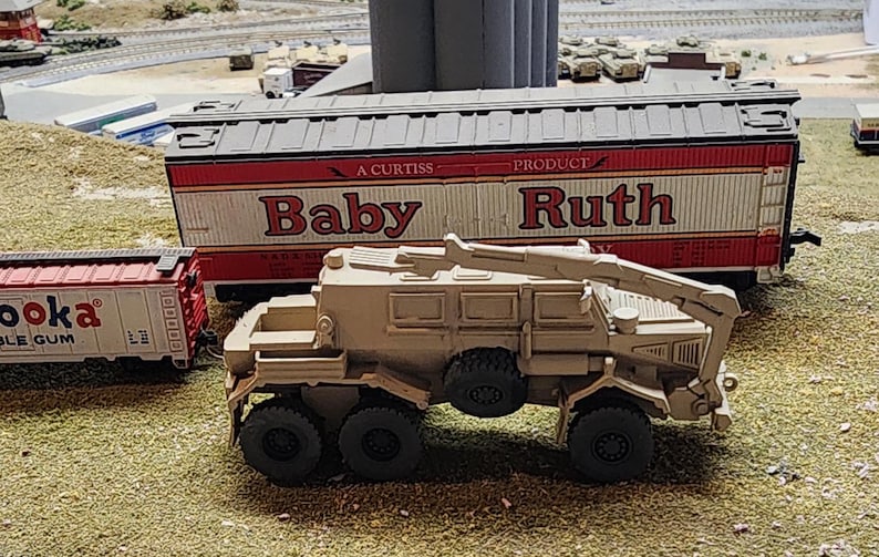 May include: A tan toy military vehicle with a crane arm, a red and white toy boxcar with the text "A Curtiss Product Baby Ruth" on the side, and a red toy boxcar with the text "Booka Bubble Gum" on the side.