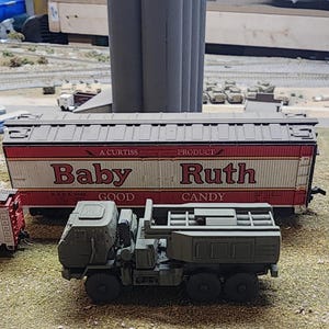 May include: A model train car with a red, white, and brown exterior. The car is labeled "A Curtiss Product Baby Ruth Candy". The car is sitting on a model train track with a green, brown, and gray background.
