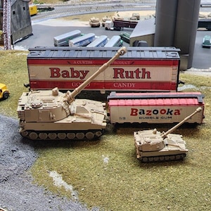 May include: Two toy military vehicles, a self-propelled howitzer and a smaller tank, are displayed on a green grassy surface. The howitzer is positioned in front of a red and white boxcar with the text "Baby Ruth Candy" printed on the side. The smaller tank is in front of a red boxcar with the text "Bazooka Bubble Gum" printed on the side.