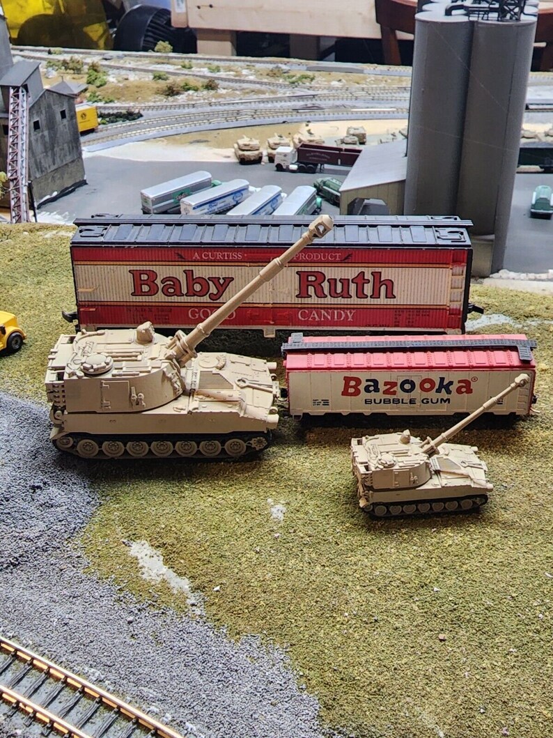 May include: Two toy military vehicles, a self-propelled howitzer and a smaller tank, are displayed on a green grassy surface. The howitzer is positioned in front of a red and white boxcar with the text "Baby Ruth Candy" printed on the side. The smaller tank is in front of a red boxcar with the text "Bazooka Bubble Gum" printed on the side.