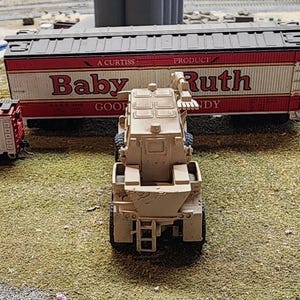May include: A tan toy military vehicle with a crane arm. The vehicle is in front of a toy train car with the words "Baby Ruth" printed on the side.