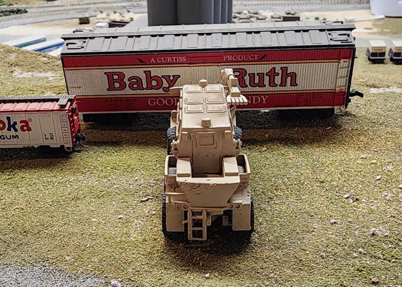 May include: A tan toy military vehicle with a crane arm. The vehicle is in front of a toy train car with the words "Baby Ruth" printed on the side.