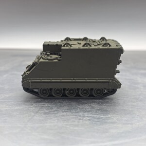 May include: A small olive green toy tank. The model tank has a rectangular body with wheels on each side and a detailed top. The toy is a replica of a military vehicle.