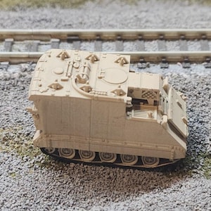 May include: A tan-colored scale model of a tracked armored personnel carrier. The vehicle features a boxy hull, a flat roof with molded hatches and equipment details, and a continuous track system along the lower sides, resting on a gravel surface.