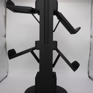 Pode incluir: Um suporte de pl&aacute;stico preto com quatro bra&ccedil;os ajust&aacute;veis para segurar fones de ouvido. O suporte tem uma base redonda.