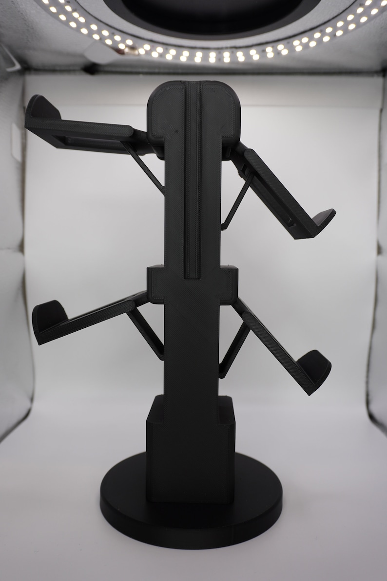 Pode incluir: Um suporte de pl&aacute;stico preto com quatro bra&ccedil;os ajust&aacute;veis para segurar fones de ouvido. O suporte tem uma base redonda.