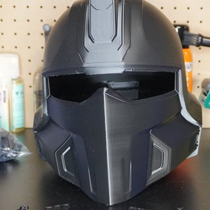 May include: A black plastic helmet with a raised design on the top. The helmet has a visor and a chin guard. There are several small black plastic pieces on the table in front of the helmet.