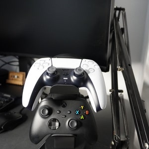Pode incluir: Um controlador de videogame preto e branco est&aacute; montado em um suporte de carregamento preto. Tamb&eacute;m h&aacute; um controlador de videogame preto no suporte. O suporte est&aacute; em uma mesa de jogo preta e vermelha.