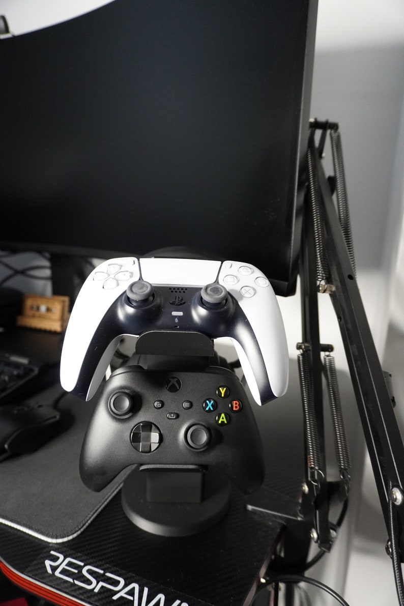 Pode incluir: Um controlador de videogame preto e branco est&aacute; montado em um suporte de carregamento preto. Tamb&eacute;m h&aacute; um controlador de videogame preto no suporte. O suporte est&aacute; em uma mesa de jogo preta e vermelha.