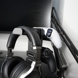 Pode incluir: Um fone de ouvido para jogos preto e prateado com um logotipo azul est&aacute; pendurado em um suporte preto com dois controladores de videogame. O suporte est&aacute; preso a um bra&ccedil;o de monitor preto.