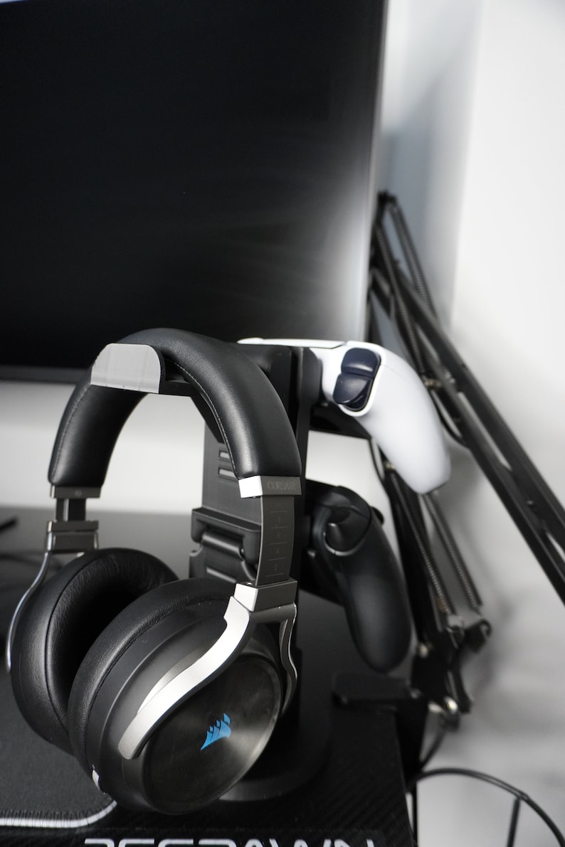 Pode incluir: Um fone de ouvido para jogos preto e prateado com um logotipo azul est&aacute; pendurado em um suporte preto com dois controladores de videogame. O suporte est&aacute; preso a um bra&ccedil;o de monitor preto.