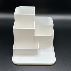 Puede incluir: Organizador de escritorio de plástico blanco con varios compartimentos. El organizador tiene una base cuadrada y un diseño escalonado con cuatro compartimentos.