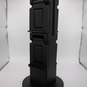 May include: A black, three-tiered, foldable phone stand with a round base. The stand is made of plastic and has a smooth, matte finish.