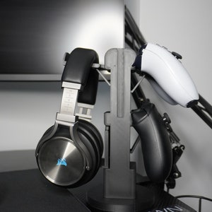 Pode incluir: Um suporte preto com um fone de ouvido prateado e preto e dois controladores pretos.