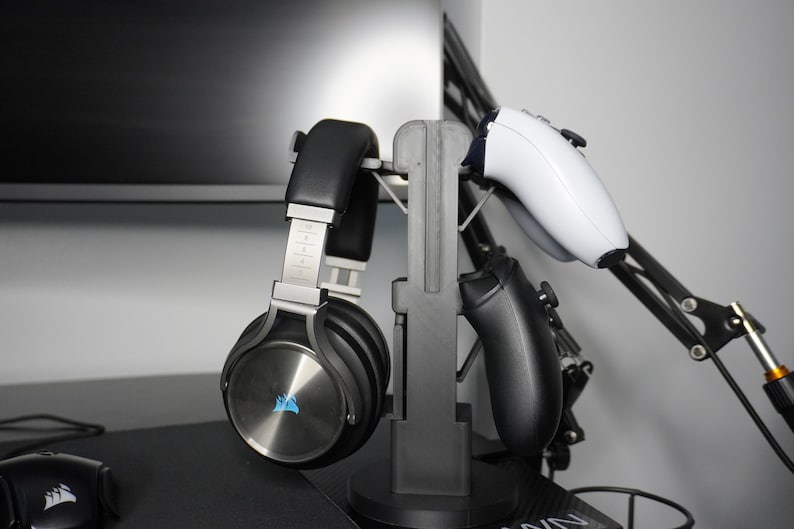 Pode incluir: Um suporte preto com um fone de ouvido prateado e preto e dois controladores pretos.