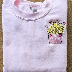 Op de afbeelding: Roze trui met een geborduurd ontwerp van een bakje friet met hartjes en de tekst "Fries Before Guys".