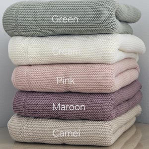 May include: A stack of five knitted blankets in different colours: green, cream, pink, maroon, and camel. The blankets are folded and stacked on top of each other.