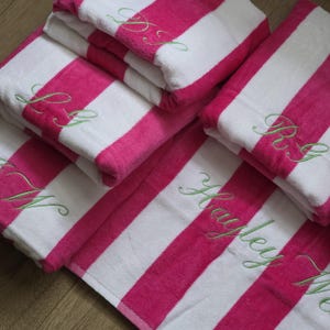 May include: A collection of folded beach towels featuring wide vertical stripes in hot pink and white. Each towel is personalised with light green cursive embroidery, including the name Hayley and various monogrammed initials on the fabric surface.