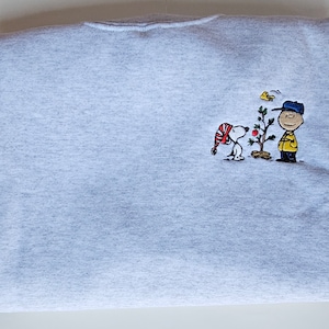 Peut inclure: Un sweat-shirt gris clair avec une broderie de Snoopy portant un bonnet de Père Noël, Charlie Brown et un petit sapin de Noël. Le motif est composé de rouge, jaune, noir et marron.