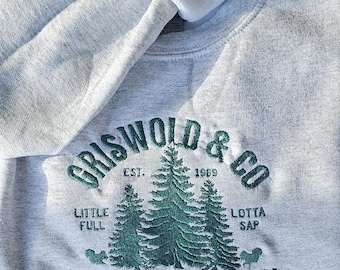 May include: A light gray sweatshirt with the embroidered text "GRISWOLD & CO CHRISTMAS TREE FARM" in green. The design includes three green Christmas trees and the phrase "a real beaut guaranteed".
