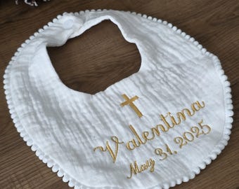 May include: White baby bib with a gold cross and embroidered text reading "Valentina May 31, 2025." The bib has a pom-pom trim and a snap closure. The fabric has a textured, crinkled appearance.