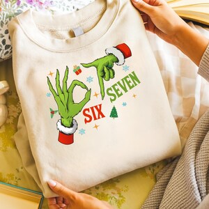 May include: Cream-coloured sweatshirt featuring a green hand with a Santa hat and the words "SIX SEVEN". The design includes snowflakes, a Christmas tree, and gift box graphics. The sweatshirt is held up by a person.