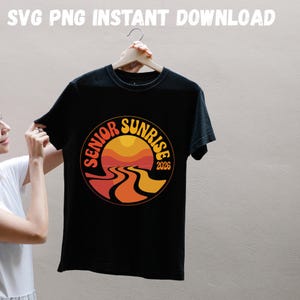 May include: Black t-shirt with a circular graphic featuring a sunset design in orange and yellow hues, with the words "Senior Sunrise 2026". The shirt is hanging on a wooden hanger.