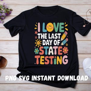 Op de afbeelding: Zwart T-shirt met een kleurrijke afbeelding waarop staat "Ik hou van de laatste dag van de staatsexamens". De afbeelding bevat een smiley, sterren, potloden, bloemen en zonnen.