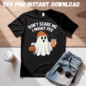 Könnte beinhalten: Schwarzes T-Shirt mit dem Text "DON'T SCARE ME I MIGHT PEE" und einem Cartoon-Geist, der einen Kürbis und ein Getränk hält. Der Geist trägt eine orangefarbene Mütze. Eine Jeans und eine Sonnenbrille liegen neben dem Shirt.