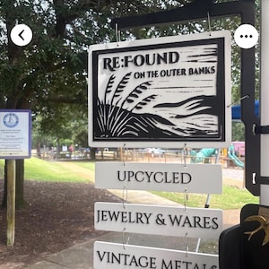 May include: A hanging sign with four white rectangular panels featuring black text and graphics. The top panel displays RE:FOUND ON THE OUTER BANKS with a coastal grass illustration. Below are three panels reading UPCYCLED, JEWELRY & WARES, and VINTAGE METALS.