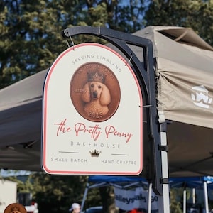May include: An arched, off-white hanging sign with a black metal bracket. The sign features a central copper-colored coin graphic with a dog wearing a crown, the text "SERVING LIMALAND", "The Pretty Penny BAKEHOUSE", and "SMALL BATCH HAND CRAFTED".
