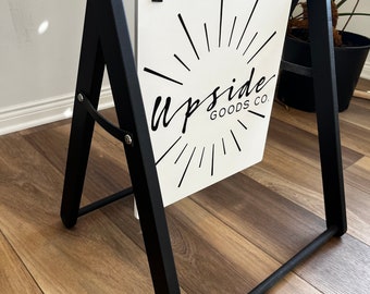 May include: A black A-frame sign stand with a white rectangular board hanging from the top bar by two black straps. The board features the black text "upside GOODS CO." centered with a black sunburst graphic radiating from the center of the text.
