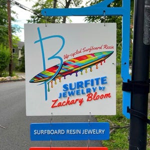 May include: A white square sign hanging from a blue bracket featuring a large blue letter B, a colorful rainbow-striped surfboard graphic with dripping resin, and the text Up-cycled Surfboard Resin, SURFITE JEWELRY by Zachary Bloom, plus two smaller hanging signs reading SURFBOARD RESIN JEWELRY and GEMSTONE JEWELRY.