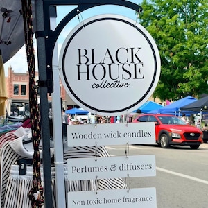 May include: A circular white sign with a black border displaying the text BLACK HOUSE collective in black font. Below the main sign, three rectangular white signs hang vertically, reading Wooden wick candles, Perfumes & diffusers, and Non toxic home fragrance.