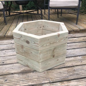 May include: A light-colored, hexagonal wooden planter box. The planter is constructed from horizontal wooden planks and has a natural wood grain finish. The planter is designed for outdoor use and is suitable for various plants.