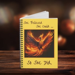 May include: A yellow notebook with a spiral binding. The cover features a phoenix rising from flames with the text "She Believed She Could... So She Did."