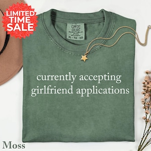 May include: A moss green t-shirt with the text "currently accepting girlfriend applications" in white. A gold star pendant necklace rests on the shirt. The shirt has a Comfort Colors tag and a "LIMITED TIME SALE" sticker.