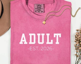 May include: A pink short-sleeved t-shirt with the white text ADULT -EST. 2026- printed on the front. The shirt features a Comfort Colors label at the inner neckline and is accompanied by a gold-toned necklace with a star-shaped pendant.