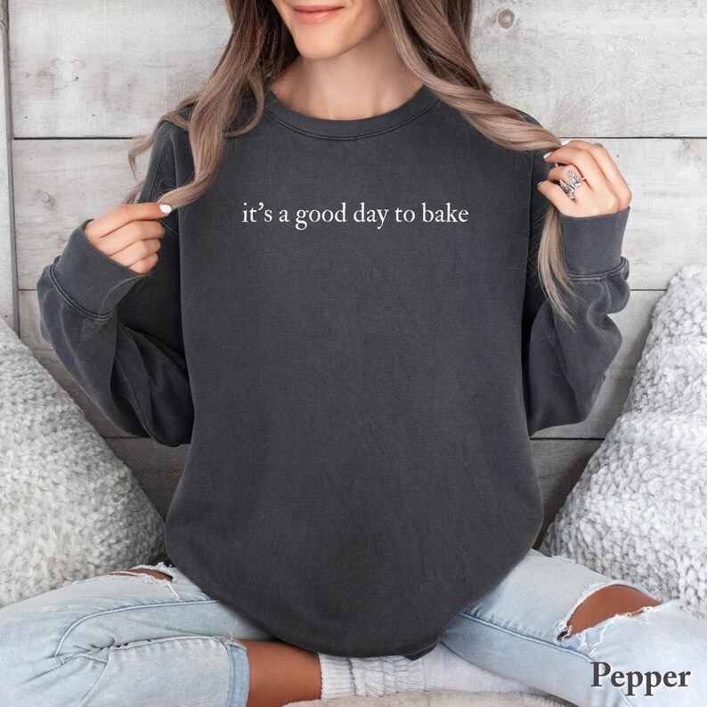 Puede incluir: Una sudadera gris oscuro con el texto blanco "it's a good day to bake". La sudadera es de cuello redondo y tiene mangas largas. La palabra "Pepper" est&aacute; impresa en blanco en la esquina inferior derecha.