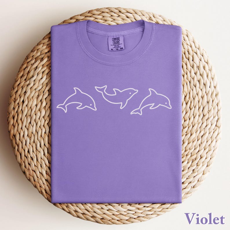 May include: A violet-colored t-shirt with a crew neck, featuring a white outline of three dolphins. The shirt is folded neatly on a woven, circular mat. The text "Comfort Colors" is visible on the tag.