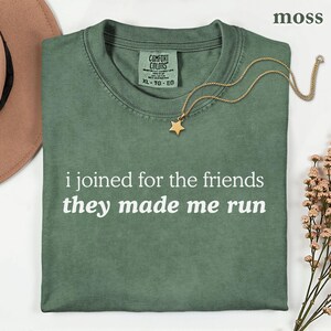 May include: A moss green crew neck t-shirt with the white text "i joined for the friends they made me run" printed on the front. The shirt features a Comfort Colors label at the inner neckline and is size XL.