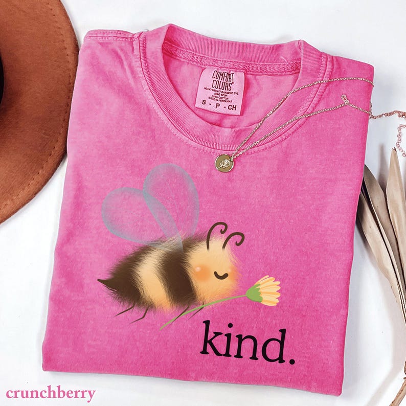 May include: A pink t-shirt with a graphic of a cartoon bee holding a flower and the word "kind." The shirt has a crew neck and short sleeves. A gold necklace is draped across the shirt. The brand name "Comfort Colors" is visible on the tag.