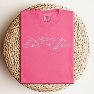 May include: A hot pink Comfort Colors t-shirt featuring a white outline of three dolphins. The shirt is folded neatly on a woven, circular placemat. The Comfort Colors label is visible at the neckline.